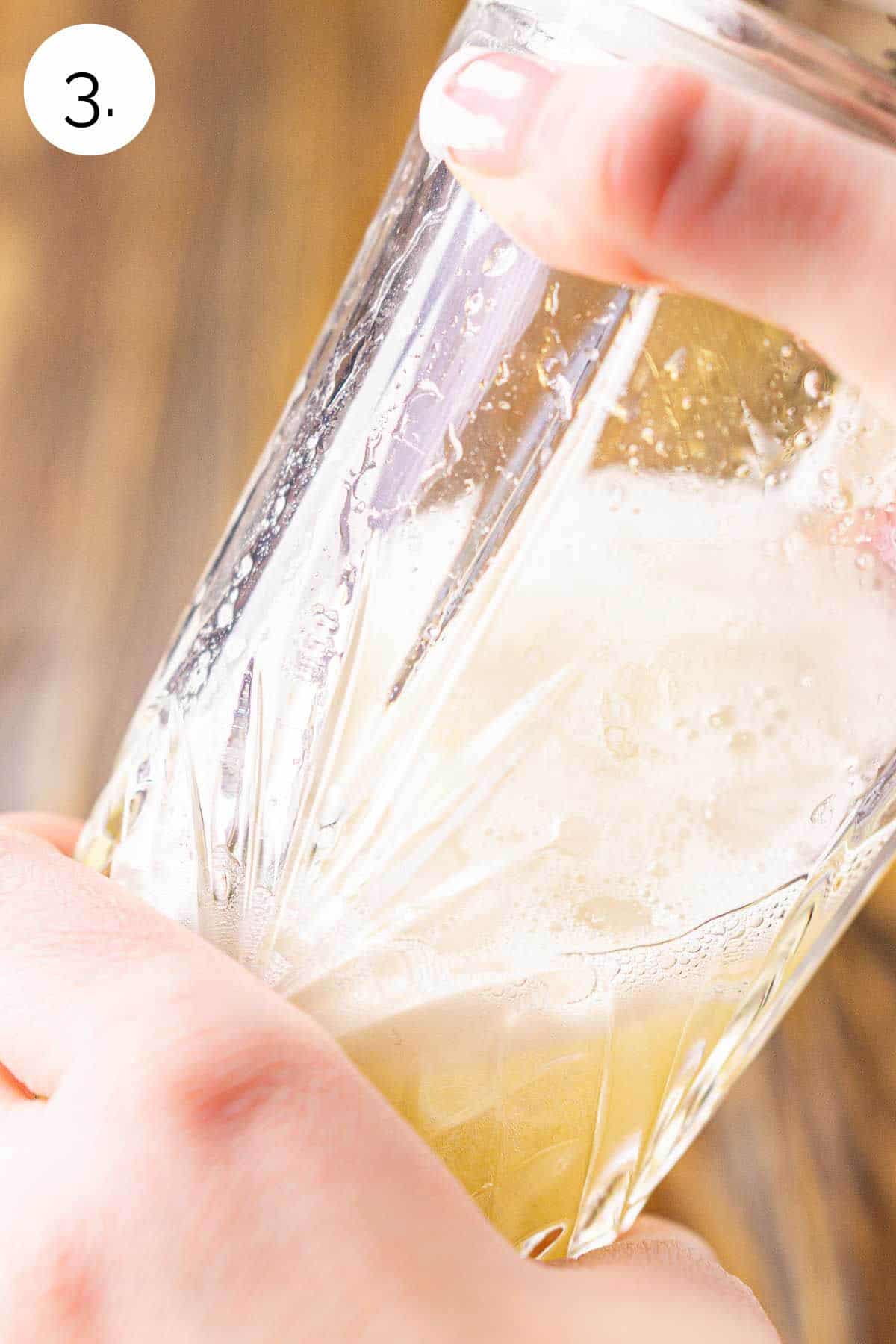 Two hands shaking the drink in a crystal cocktail shaker to mix against a brown wooden surface.