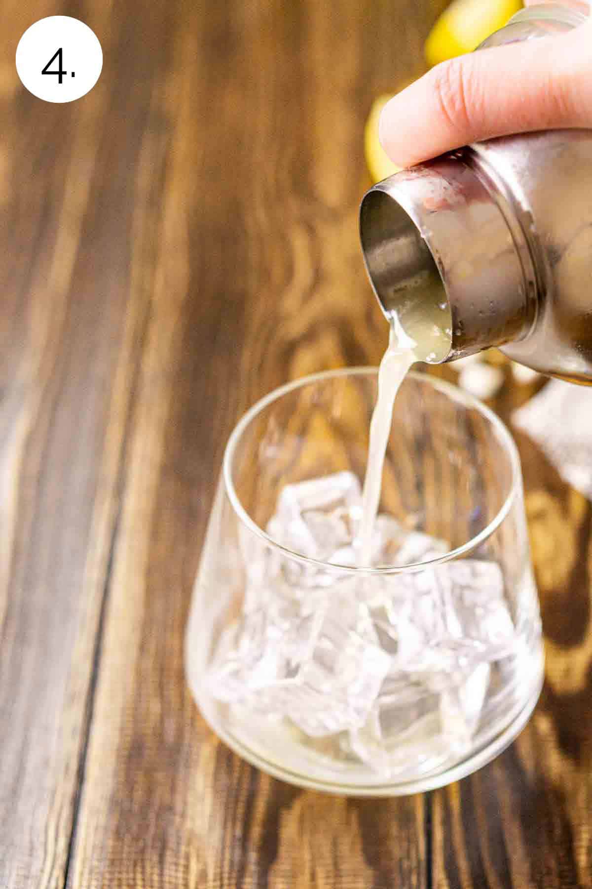 A hand holding a cocktail shaker as it strains the drink into a rocks glass filled with ice on a brown wooden surface.