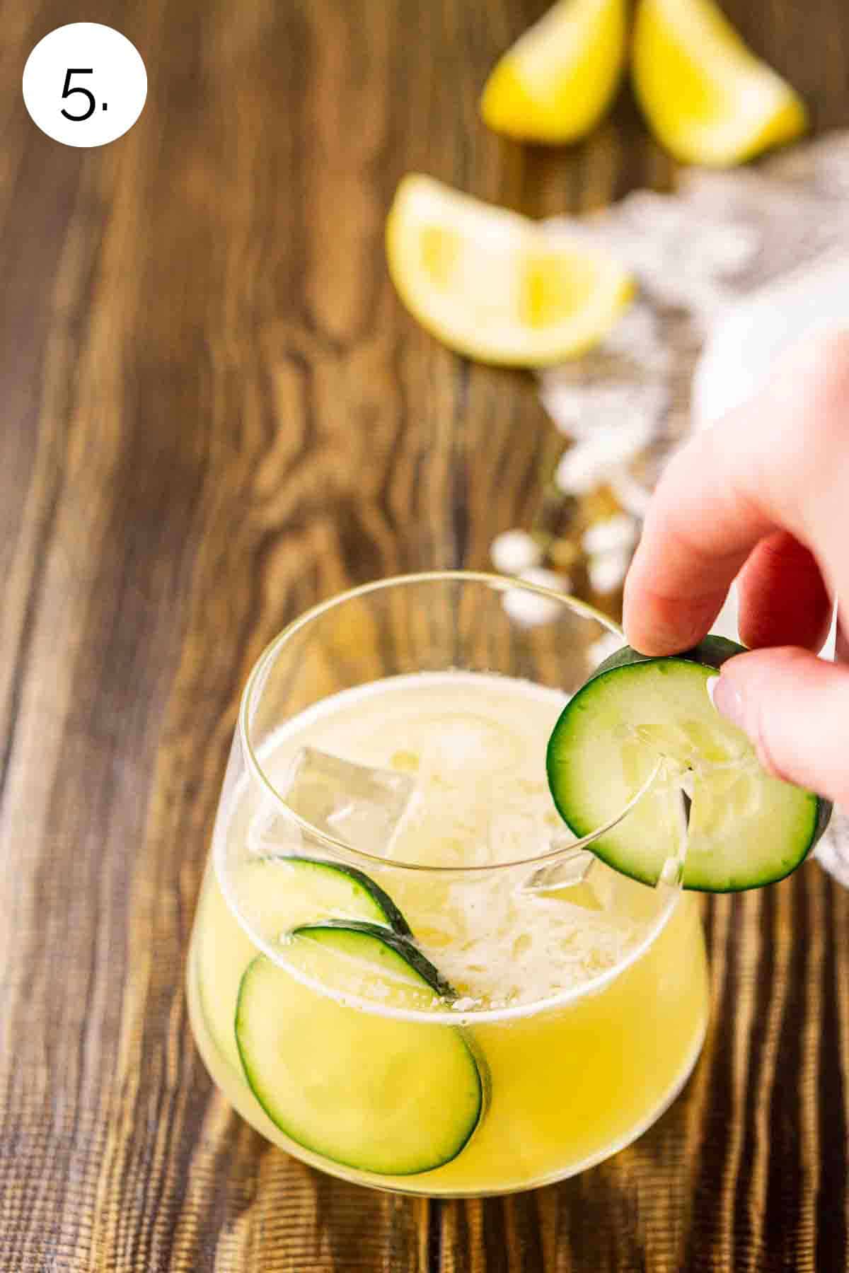 A hand placing a cucumber slice on the rim of the serving glass on a brown wooden surface.