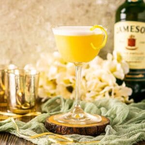 A Jameson whiskey sour on a wooden coaster on top of green cloth with a bottle of Jameson, white flowers and candles in the background.