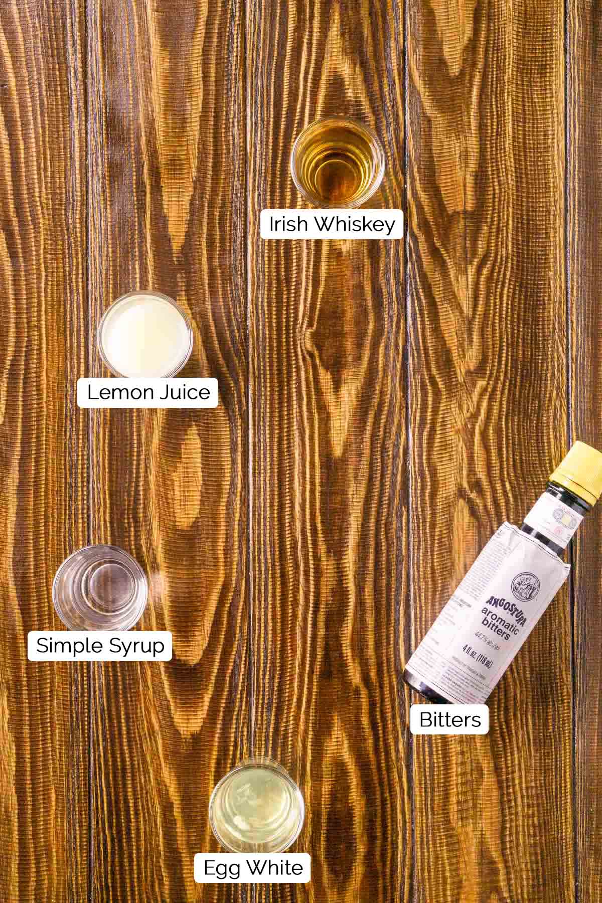 The drink ingredients in shot glasses with black and white labels underneath the items on a brown wooden surface.
