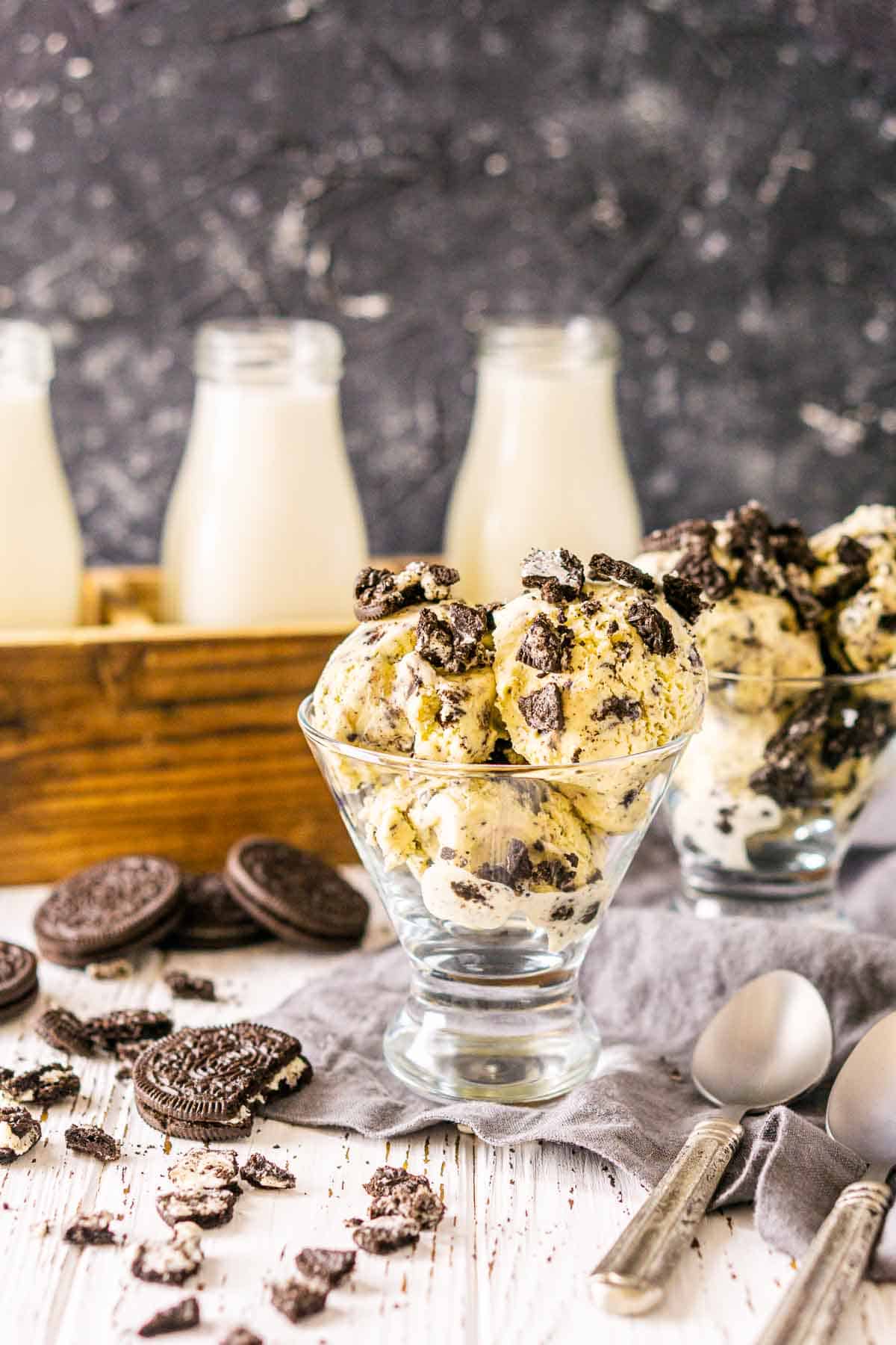 A glass cup of the Oreo ice cream on a white wooden board with silver spoons to the right and crushed cookies to the left.
