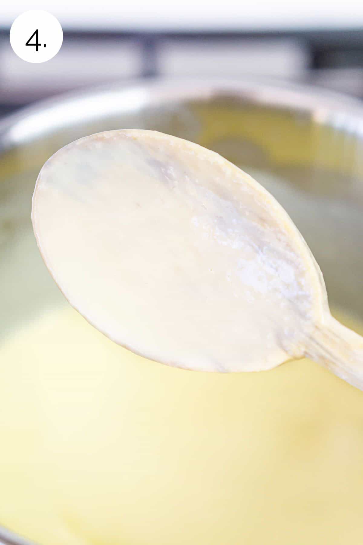 The custard mixture covering a wooden spoon after thickening in a stainless steel saucepan on the stove.