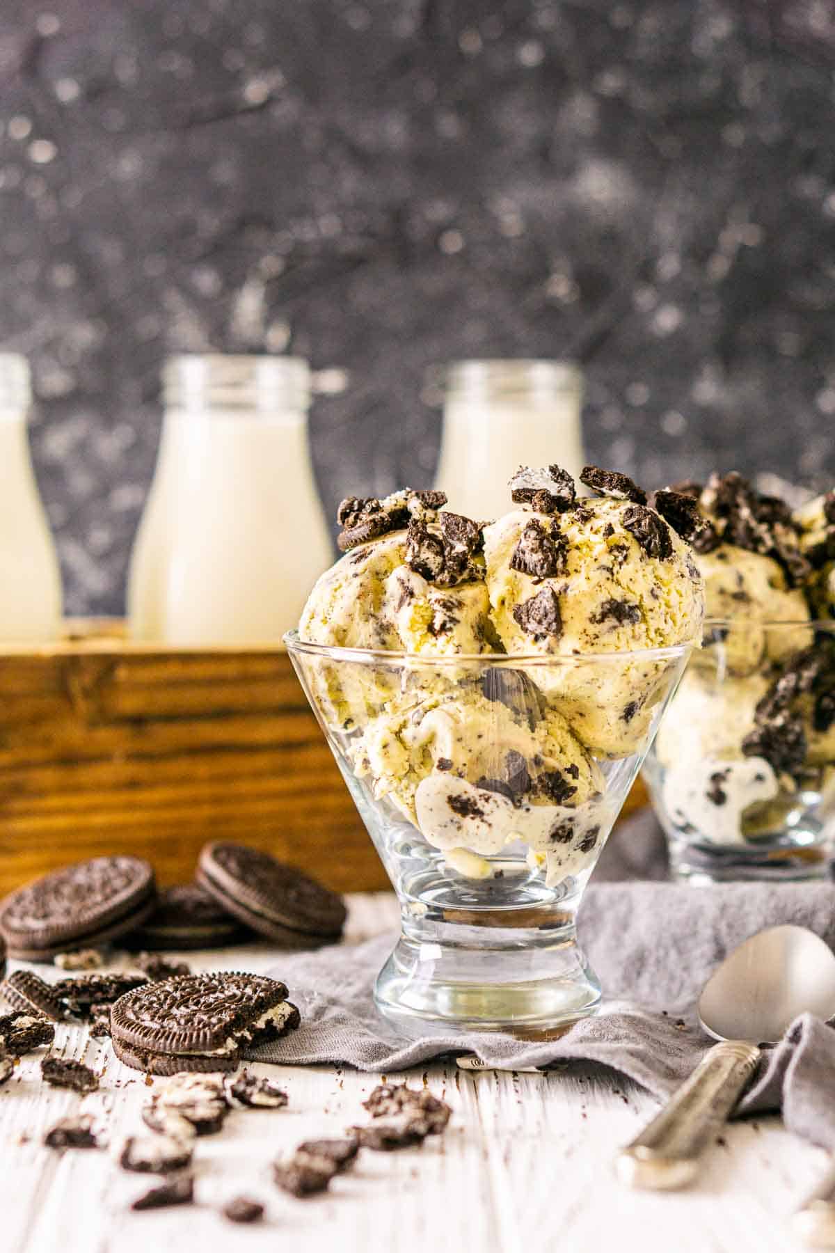 A close-up of the Oreo ice cream on a white wooden surface with milk in the background and crushed cookies around it.