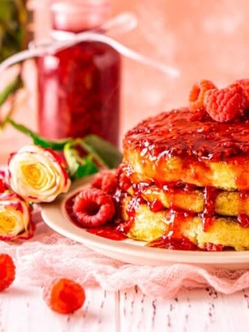 A stack of raspberry pancakes on a small plate against a pink background with pink and white flowers and berries to the side.