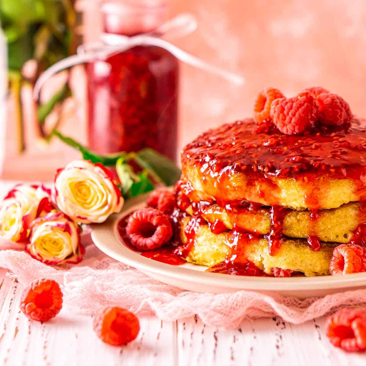 A stack of raspberry pancakes on a plate with flowers and berries around it.