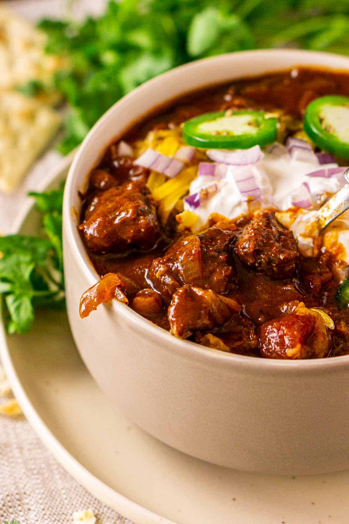 A close-up of the chuck roast in the smoked chili in a cream-colored bowl on a small plate with a bundle of cilantro in the background.