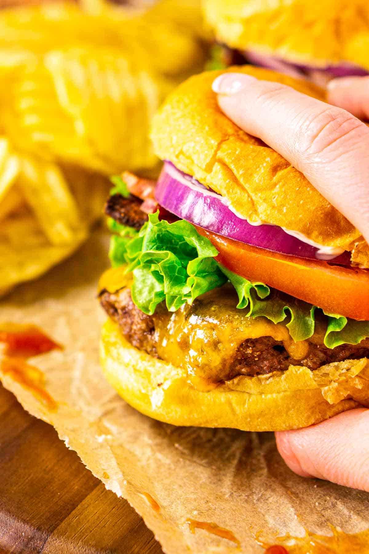 A close-up of a hand grabbing a venison burger on parchment paper and a wooden serving tray.