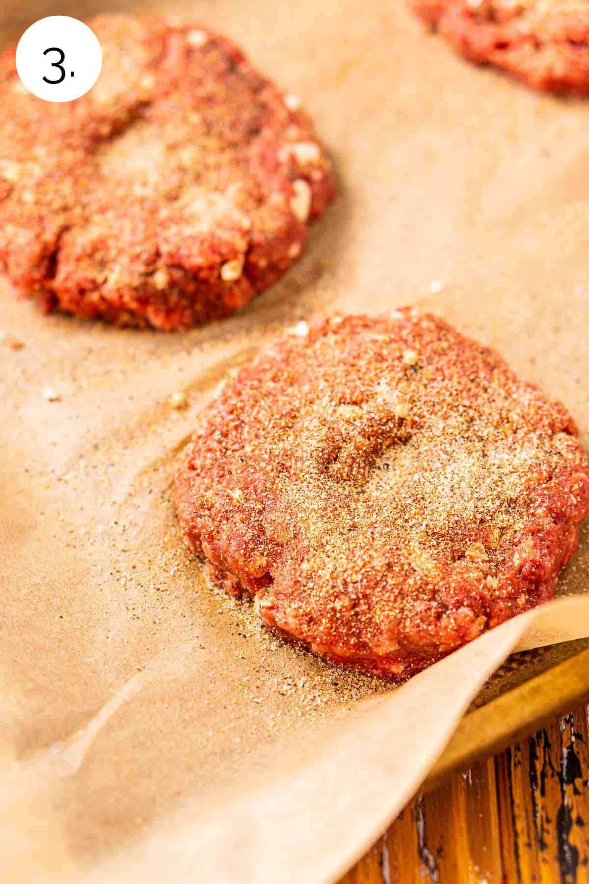 The burger patties on a parchment paper-lined baking sheet with seasoning on top before they go on the grill.