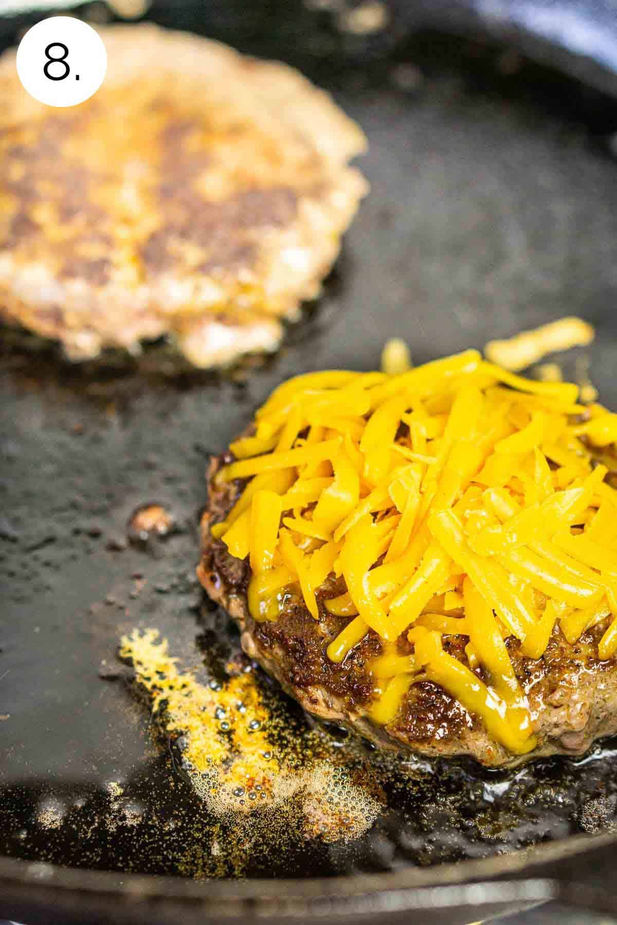 The patties in a black cast-iron skillet with shredded cheddar cheese on top as it melts.