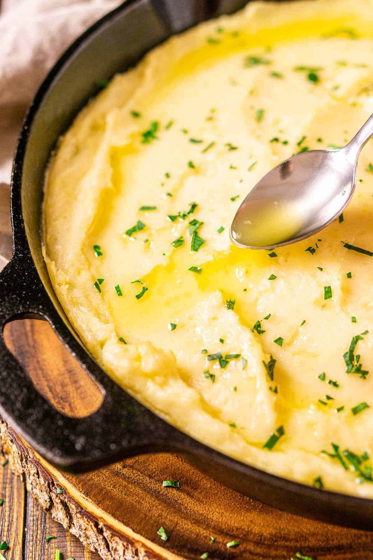 A silver spoon pouring melted butter on top of mashed potatoes in a black cast-iron skillet on top of a wooden serving platter.