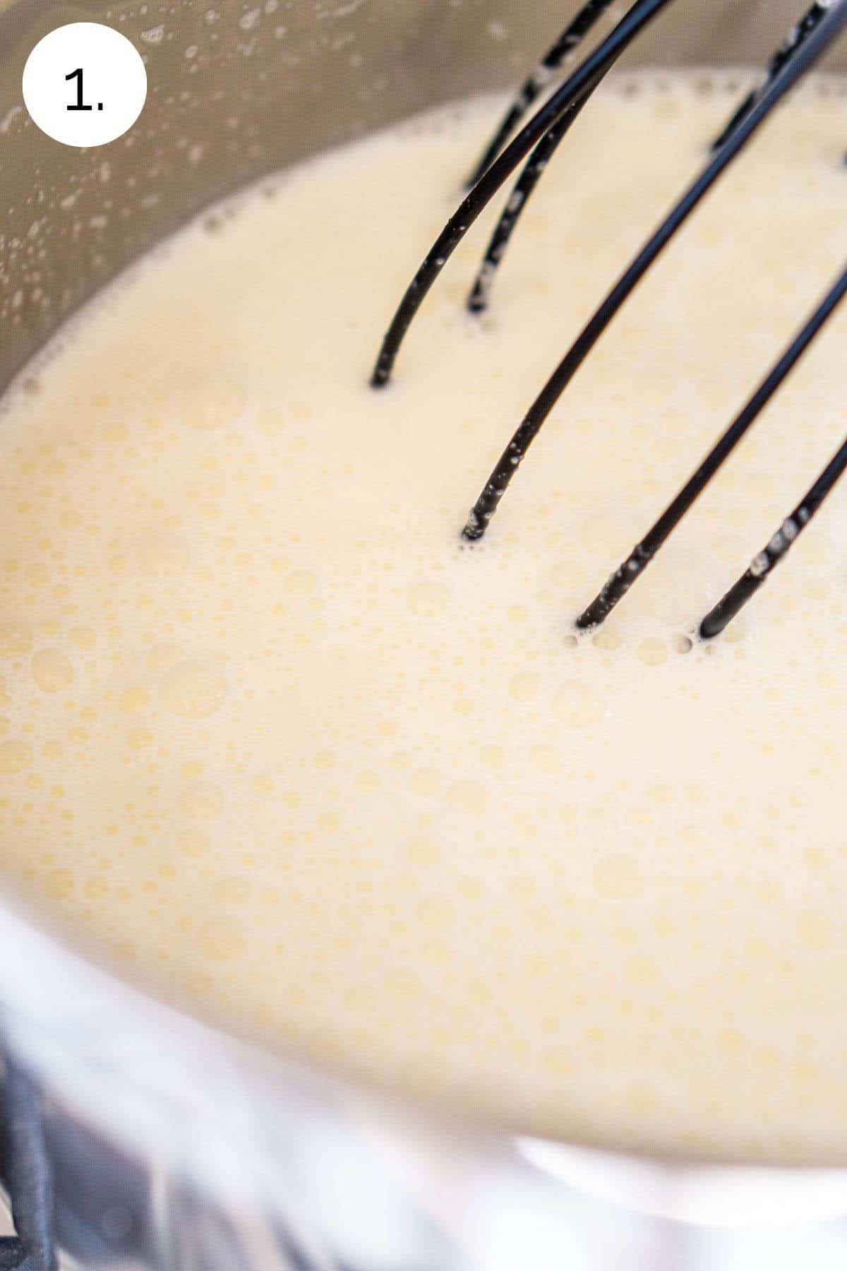 A black whisking mixing the cream, milk and sugar in a small stainless steel saucepan as it simmers on the stove.