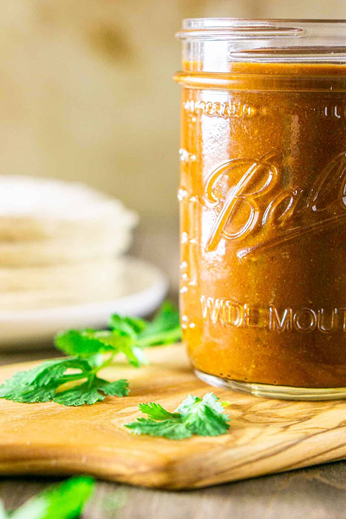 A glass mason jar filled with the red enchilada sauce on a wooden serving tray surrounded by cilantro and corn tortillas.