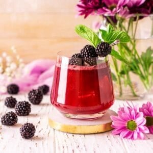 Looking straight on to a blackberry bourbon smash on a white wooden surface with purple flowers and fresh berries on the side.