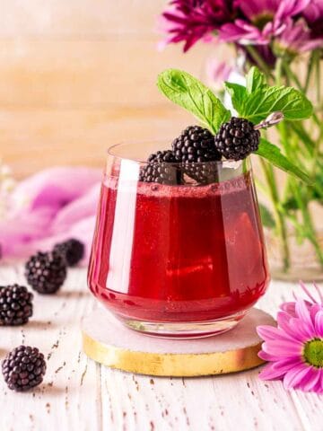 Looking straight on to a blackberry bourbon smash on a white wooden surface with purple flowers and fresh berries on the side.
