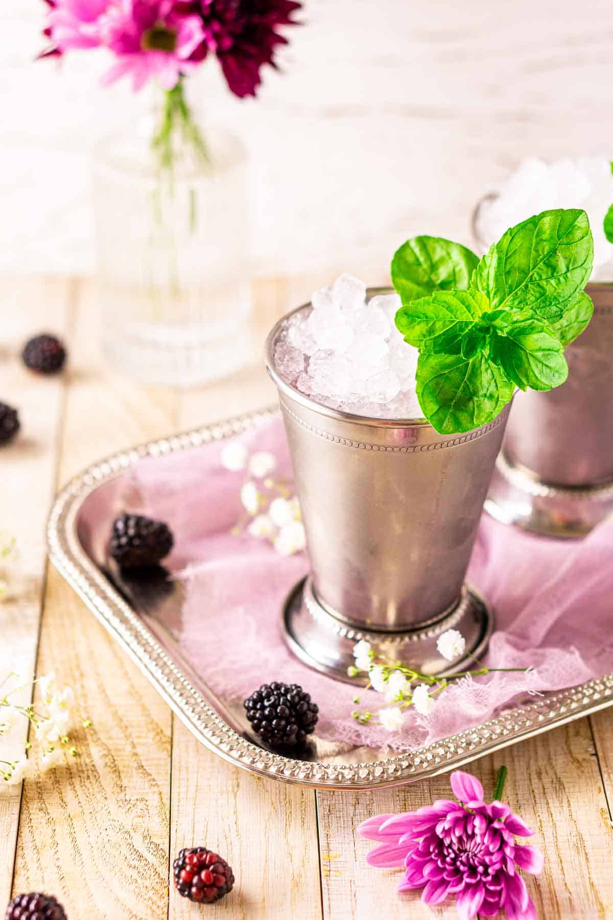 A close-up of a blackberry mint julep on a silver tray with berries and purple and white flowers around it.