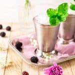 A blackberry mint julep in a silver cup on a serving tray with purple cloth and berries surrounding the drink.