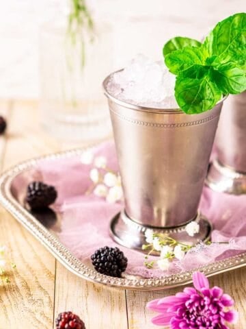 A blackberry mint julep in a silver cup on a serving tray with purple cloth and berries surrounding the drink.