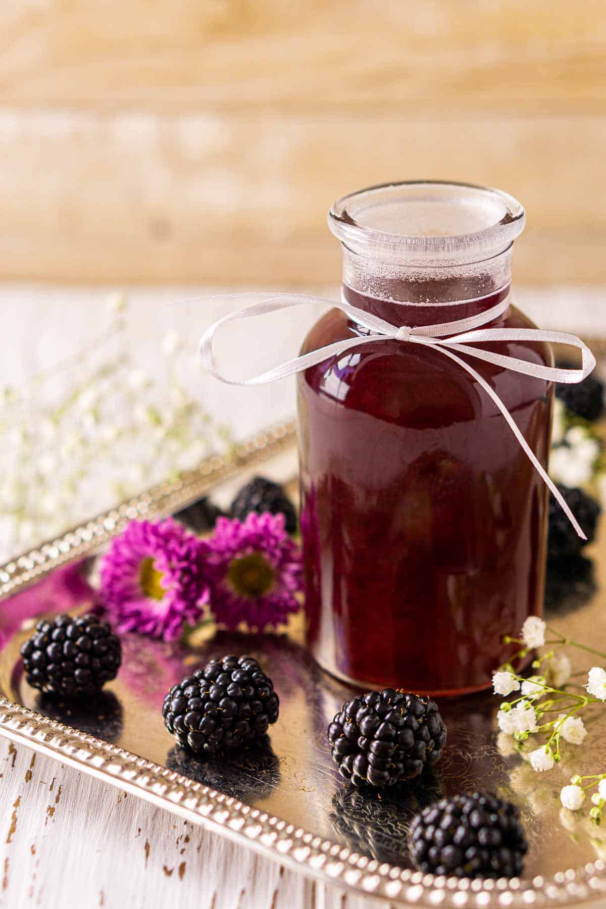 A bottle of the blackberry simple syrup on a silver tray with berries and white flowers against a cream-colored background.