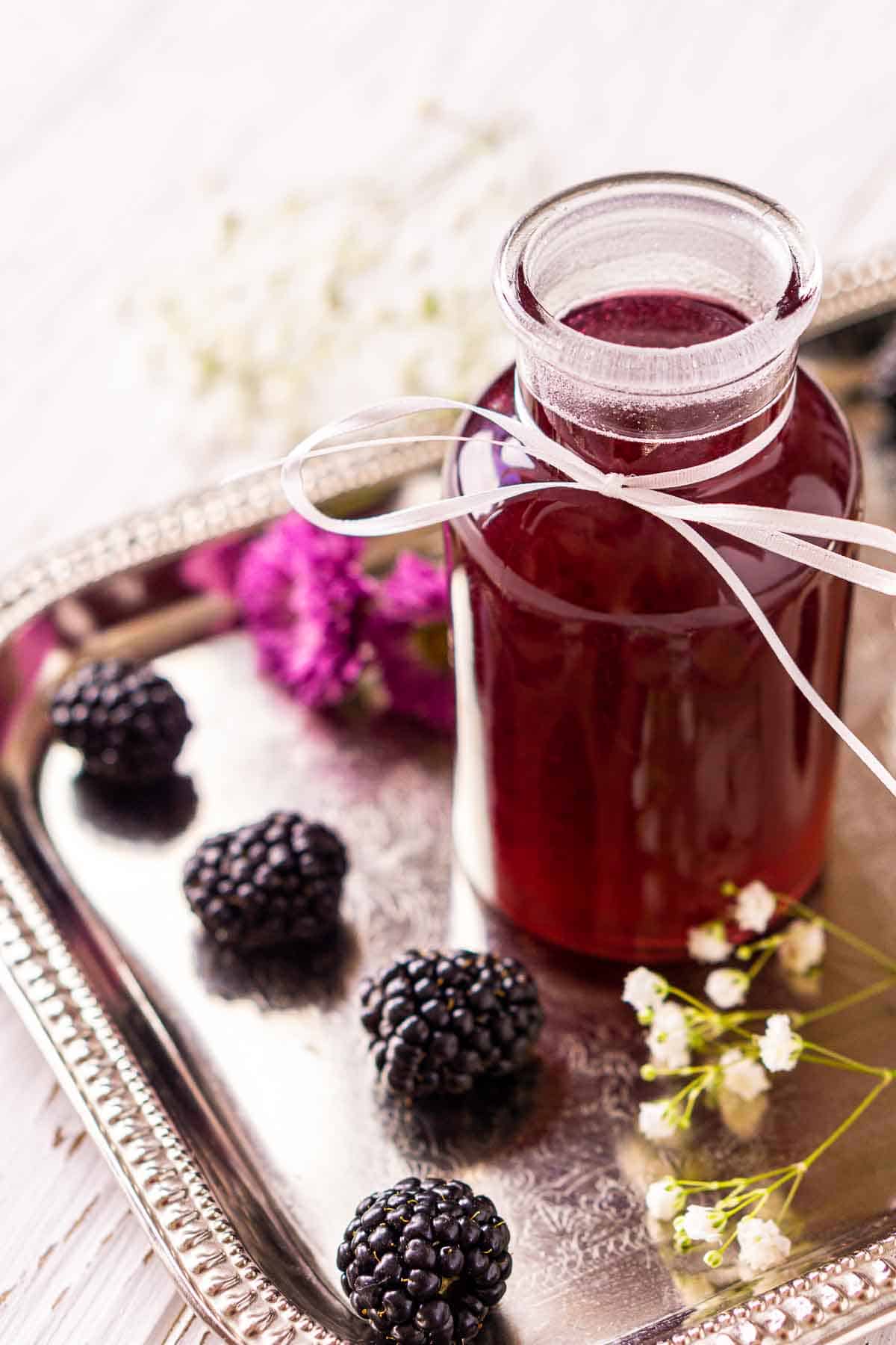A side view of the blackberry simple syrup on a silver serving tray with berries and flowers around it.