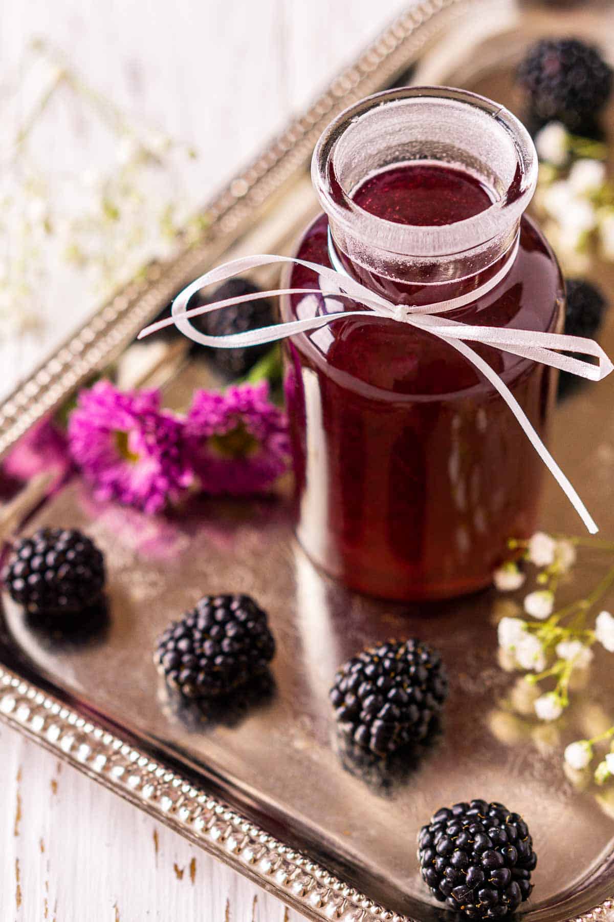 Looking down on a bottle of the blackberry simple syrup on a silver tray with fresh berries and white flowers around it.