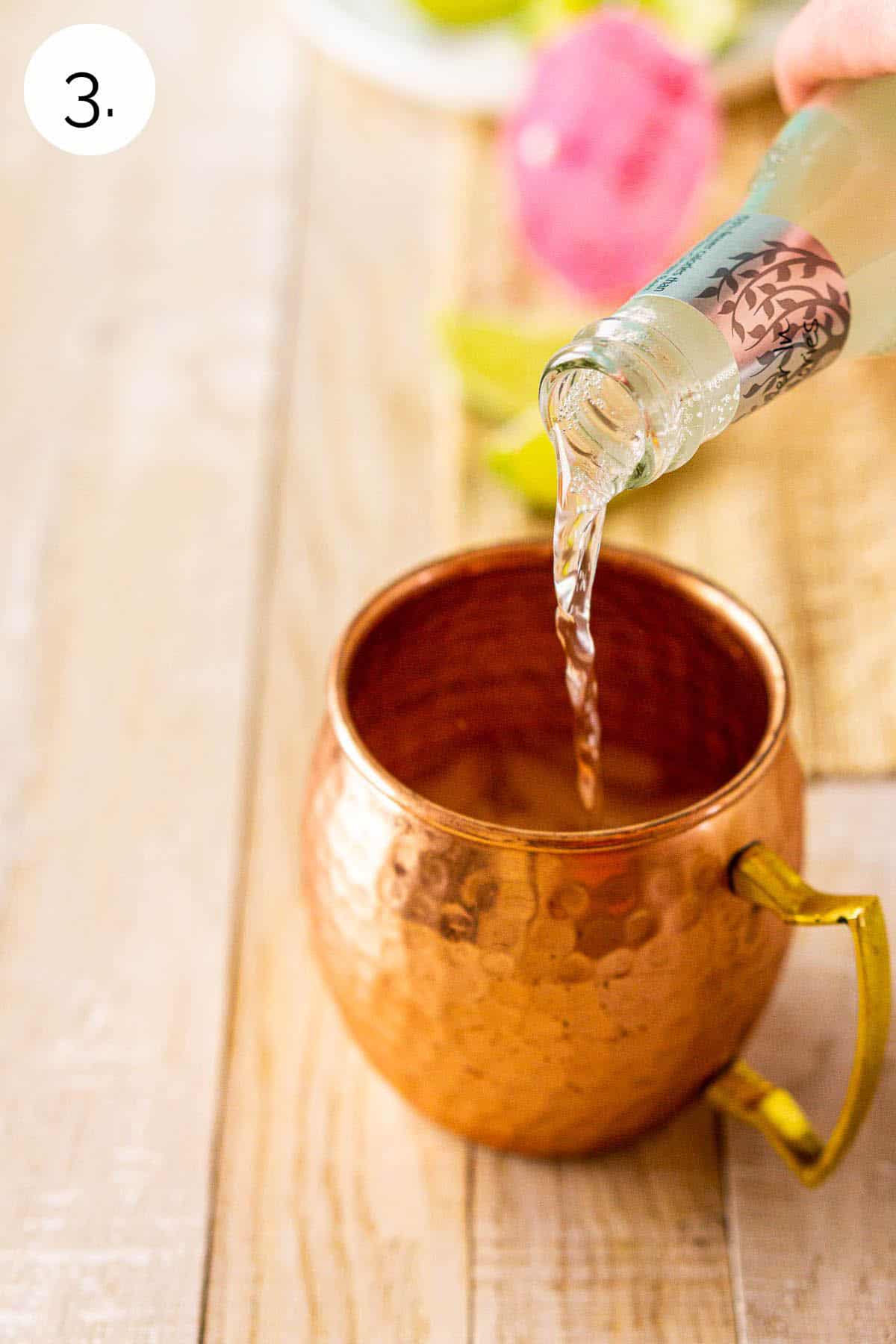 A hand pouring a bottle of ginger beer into the copper mug on a cream-colored wooden surface.