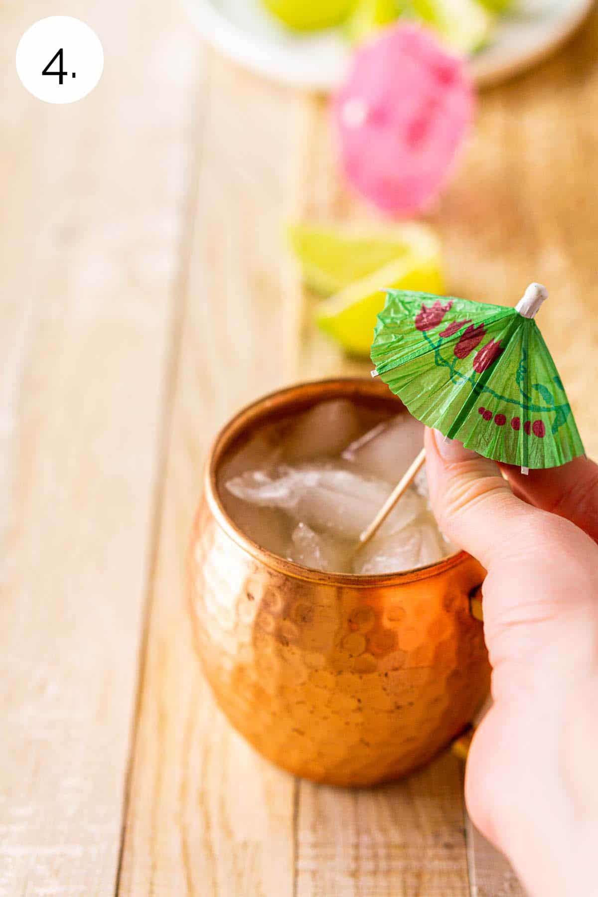 A hand placing a tiki cocktail umbrella in the copper mug on a cream-colored wooden board.
