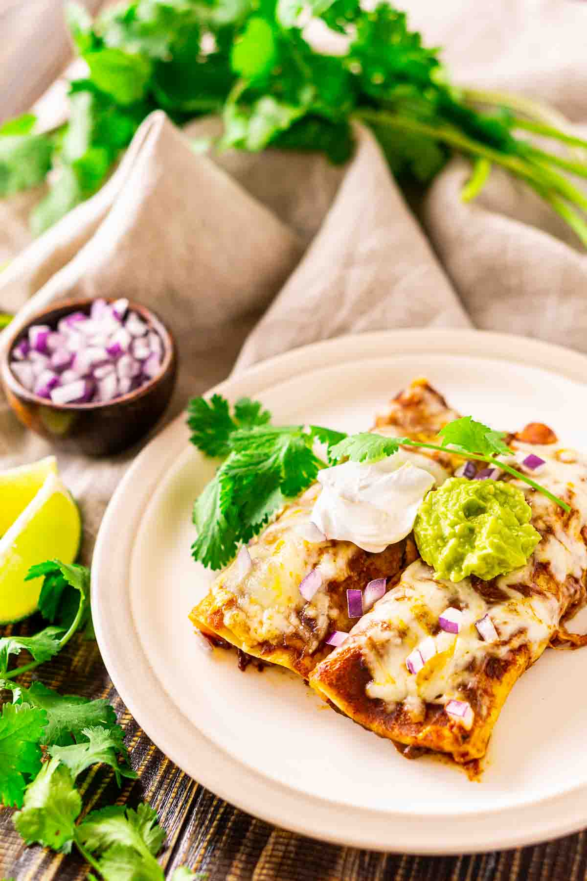 Two shredded beef enchiladas on a white plate with cilantro sprigs and lime slices to the left.