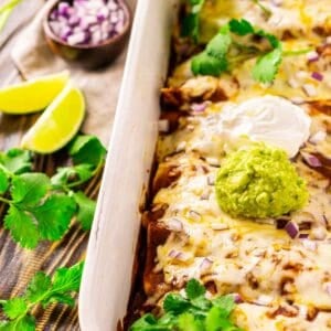 Looking down on a pan of shredded beef enchiladas on a brown wooden surface with cilantro, red onion and lime slices to the left.