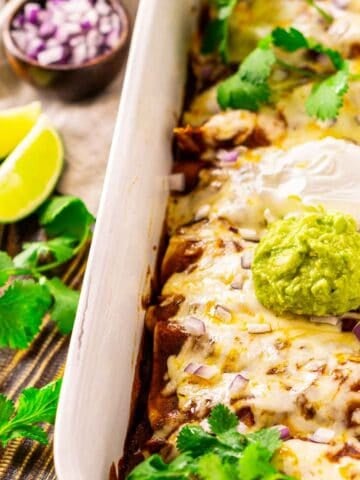 Looking down on a pan of shredded beef enchiladas on a brown wooden surface with cilantro, red onion and lime slices to the left.