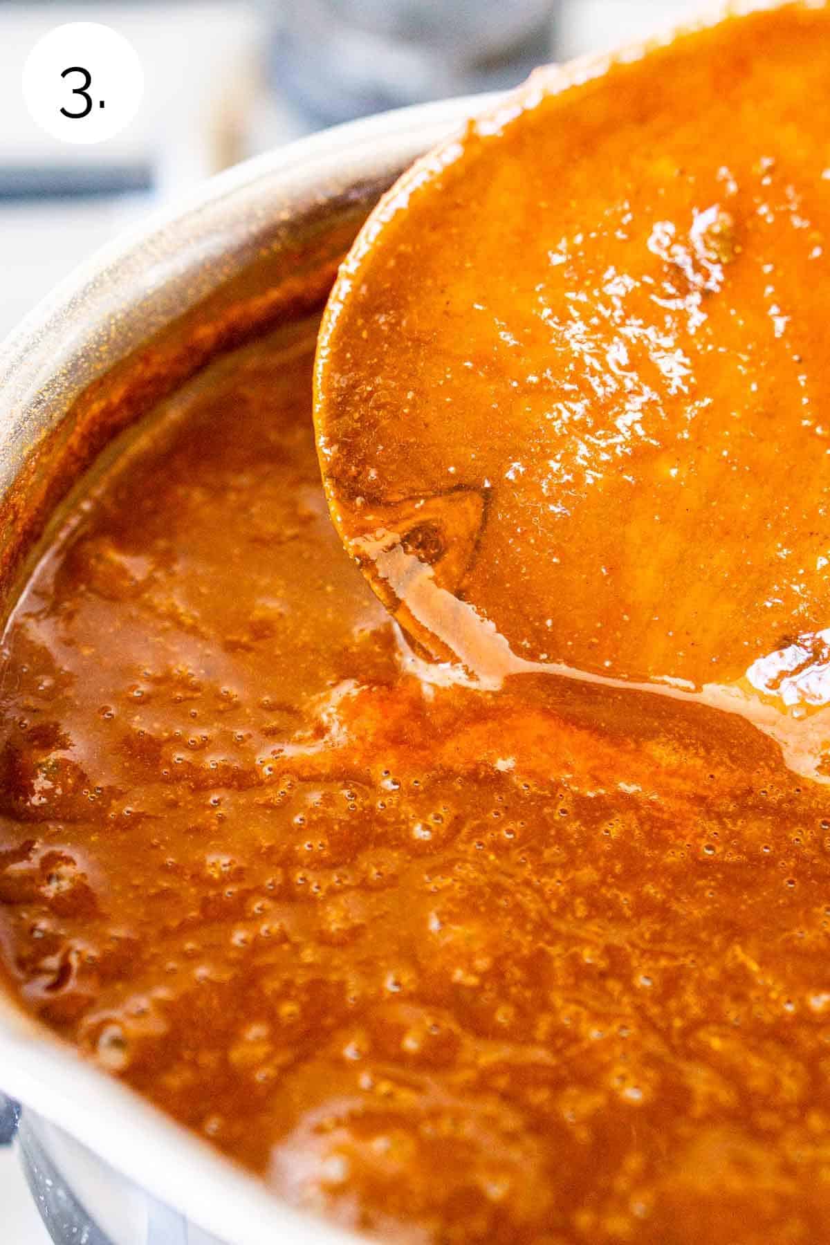 A wooden spoon covered with the enchilada sauce over the rest of the sauce in a small stainless steel saucepan.
