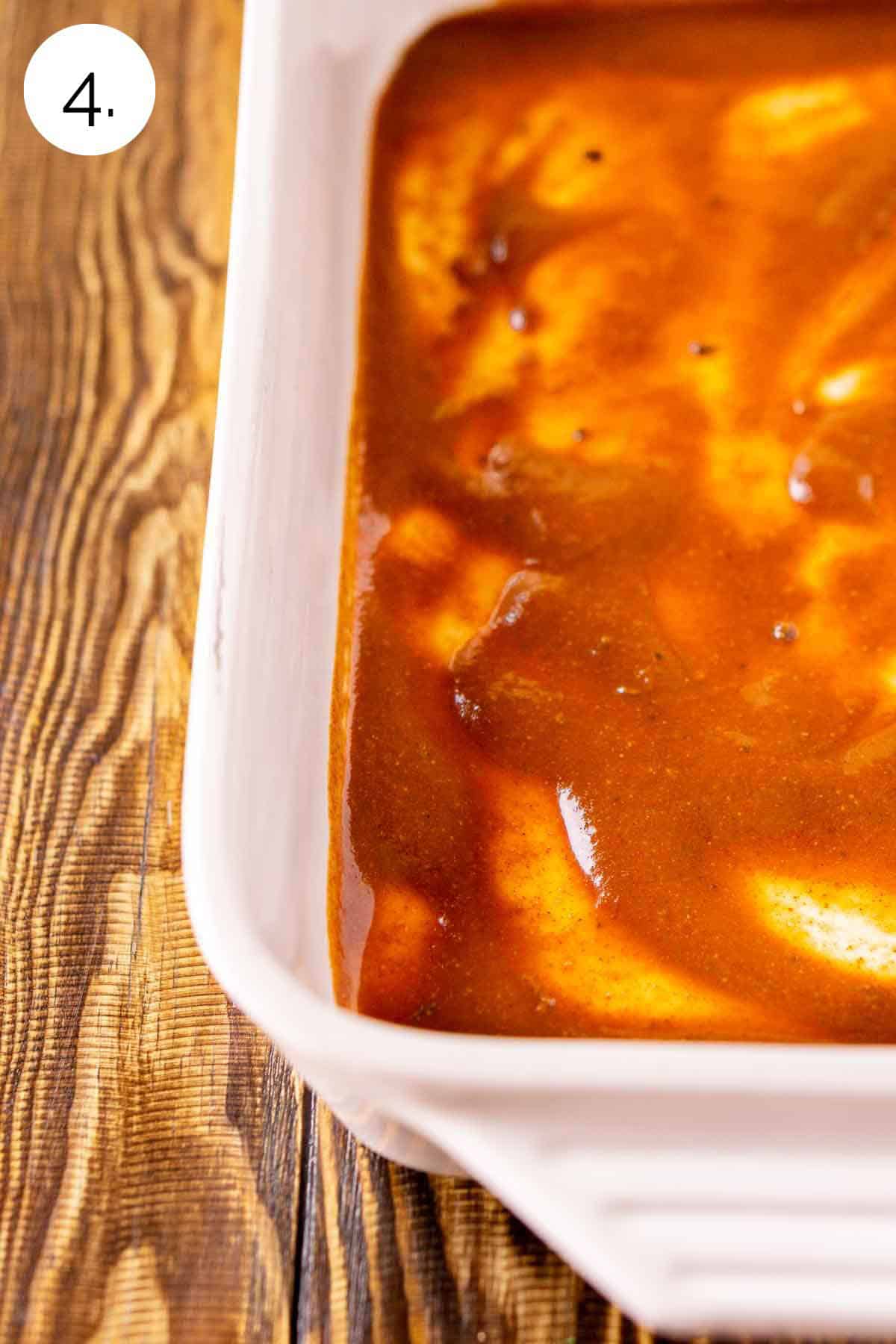 A white baking dish on a brown wooden surface with red enchilada sauce spread on the bottom of the pan.