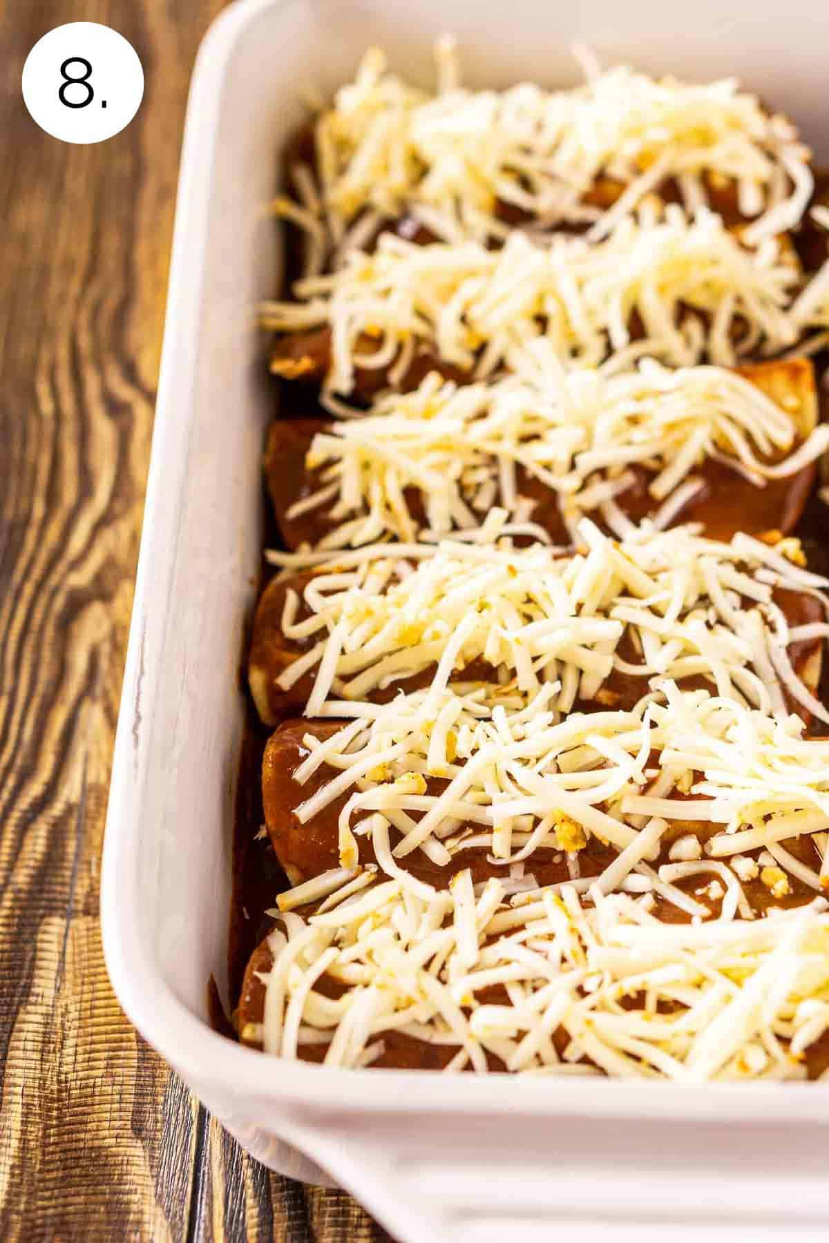 The enchiladas in a white baking dish on a brown wooden surface covered in the sauce and cheese before baking.