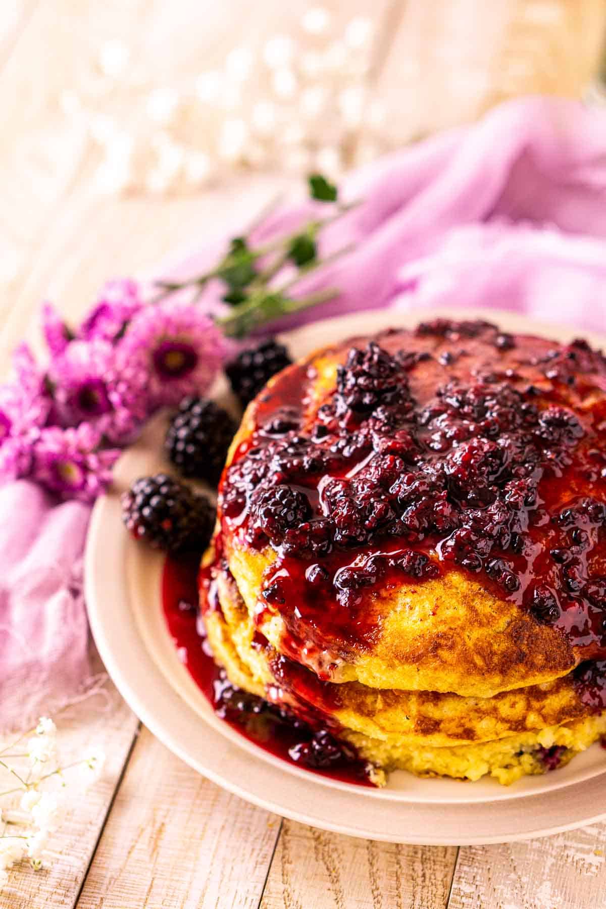 Looking down on a stack of blackberry pancakes with syrup on top and white flowers to the left and in the background.