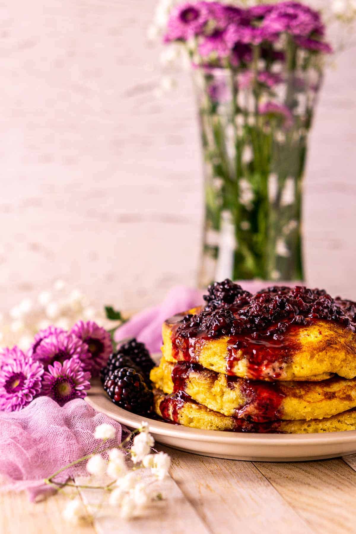 A side view of the blackberry pancakes on a cream-colored surface with purple cloth and white flowers on the left of the plate.