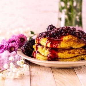 Looking straight on a stack of blackberry pancakes on a small plate with a bundle of purple and white flowers to the side.