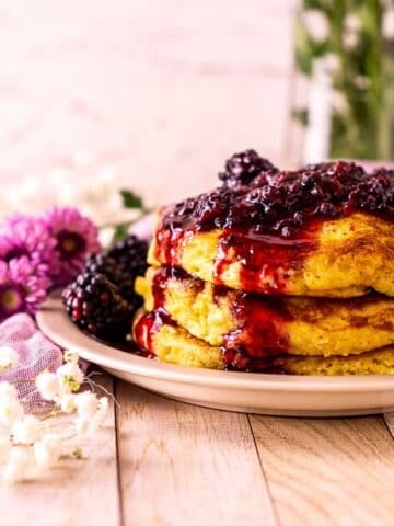 Looking straight on a stack of blackberry pancakes on a small plate with a bundle of purple and white flowers to the side.