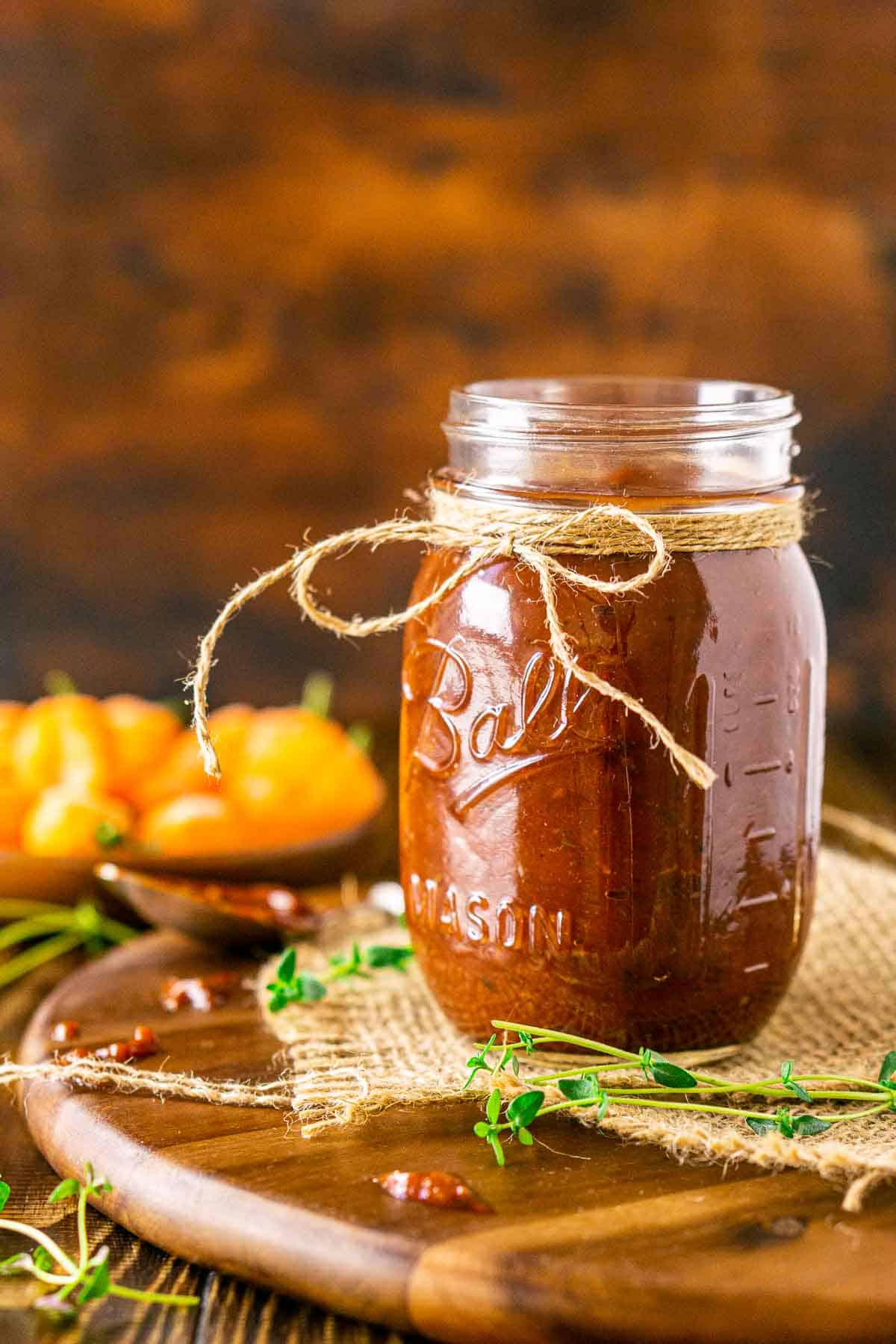 Looking straight on to a jar of jerk BBQ sauce on a wooden tray with burlap and sprigs of thyme scattered around it.