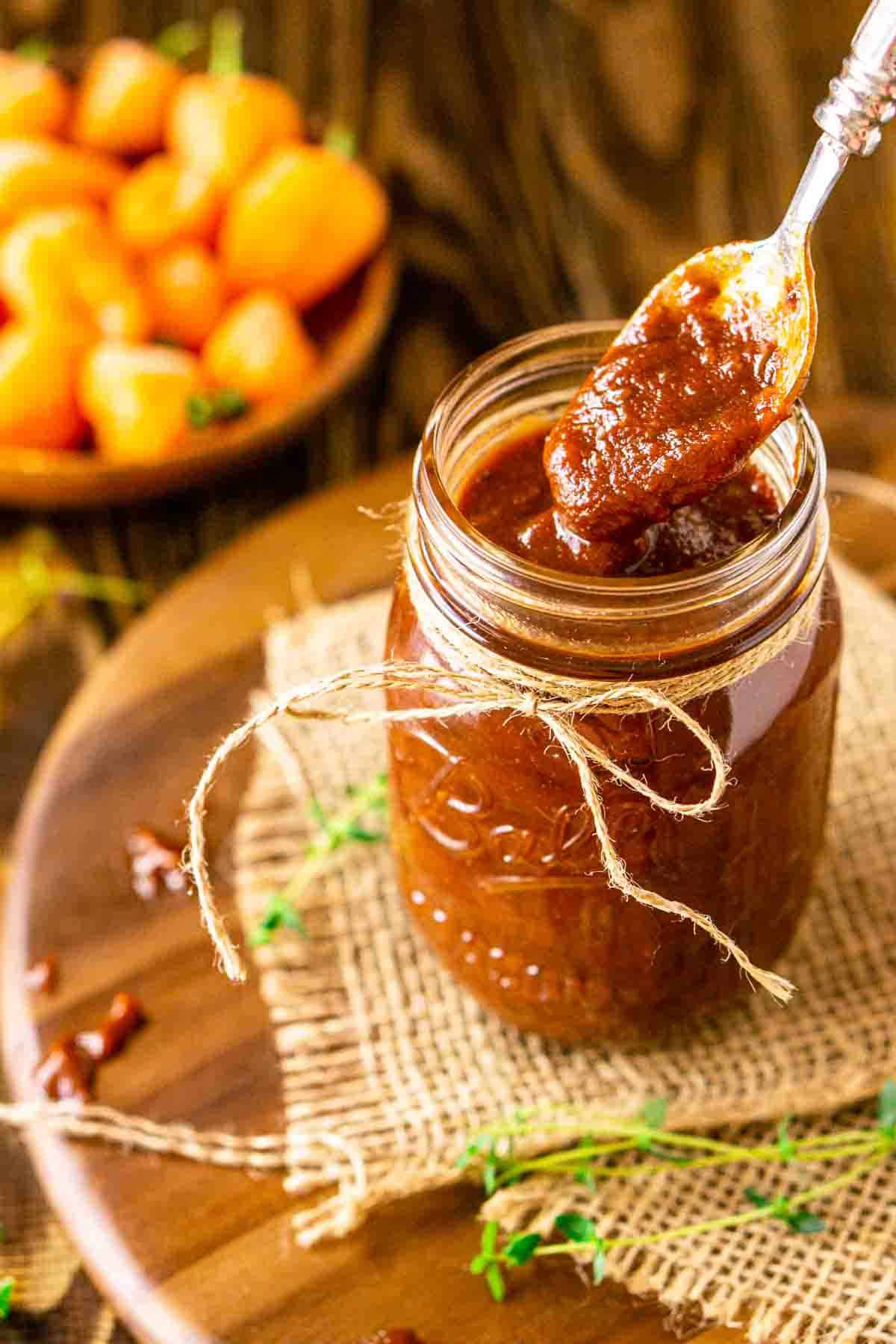 A rustic silver spoon drizzling the jerk BBQ sauce into a jar on a wooden platter with a piece of burlap and peppers in the background.