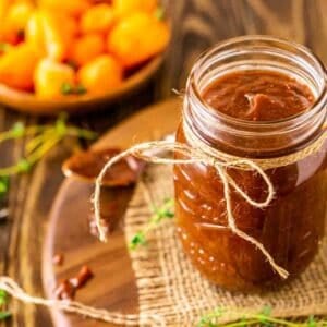 A glass mason jar filled with the jerk BBQ sauce on a wooden tray with a piece of folded burlap and fresh thyme and peppers around it.