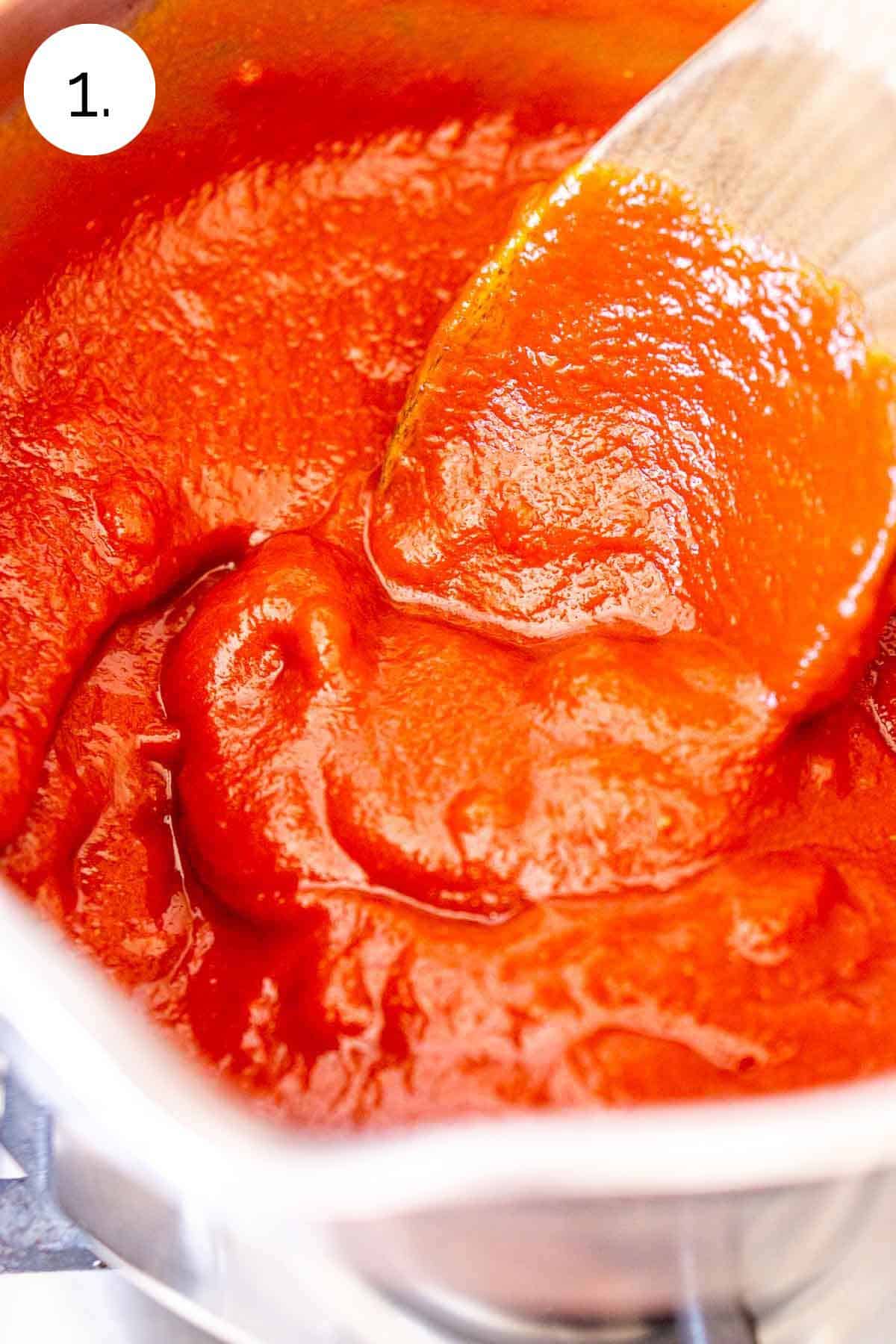 A wooden spoon stirring the tomato sauce and tomato paste in a small saucepan on the stove.