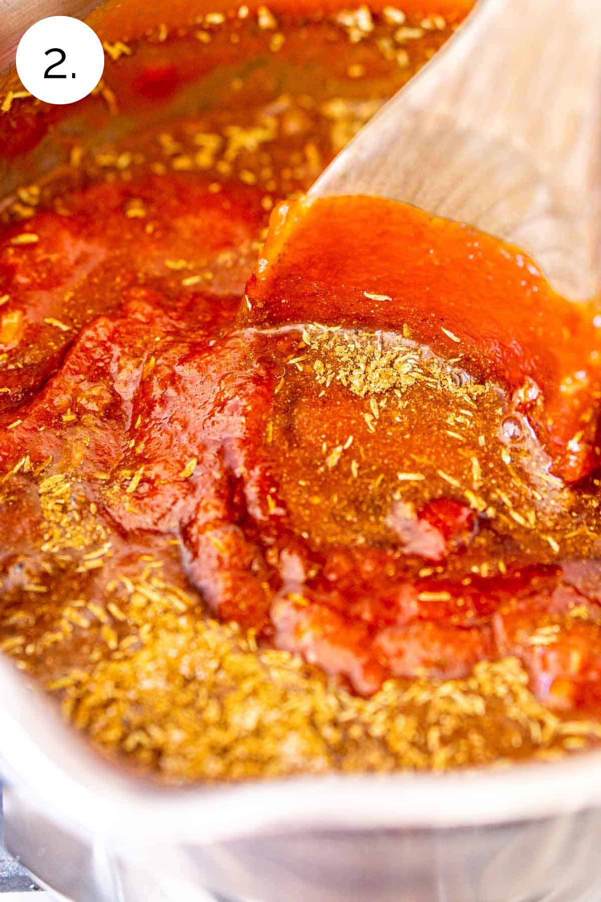 A brown wooden spoon stirring the spice mixture into the tomato sauce in a small stainless steel saucepan on the stove.