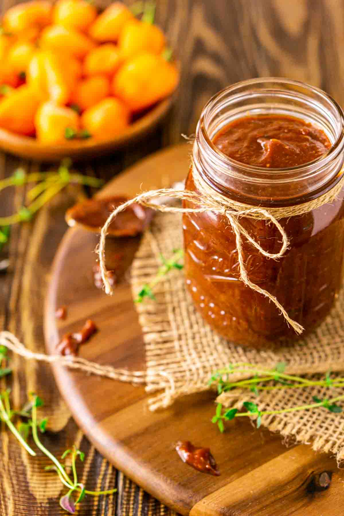 A glass mason jar filled with the jerk BBQ sauce on a wooden tray with a piece of folded burlap and fresh thyme and peppers around it.