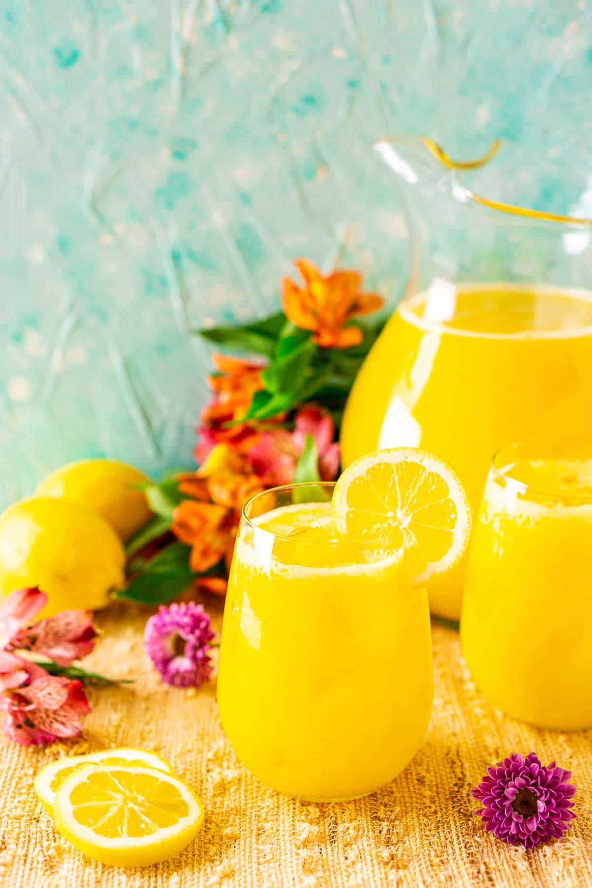 Looking down on a glass of mango lemonade with tropical flowers around it and a pitcher to the right.