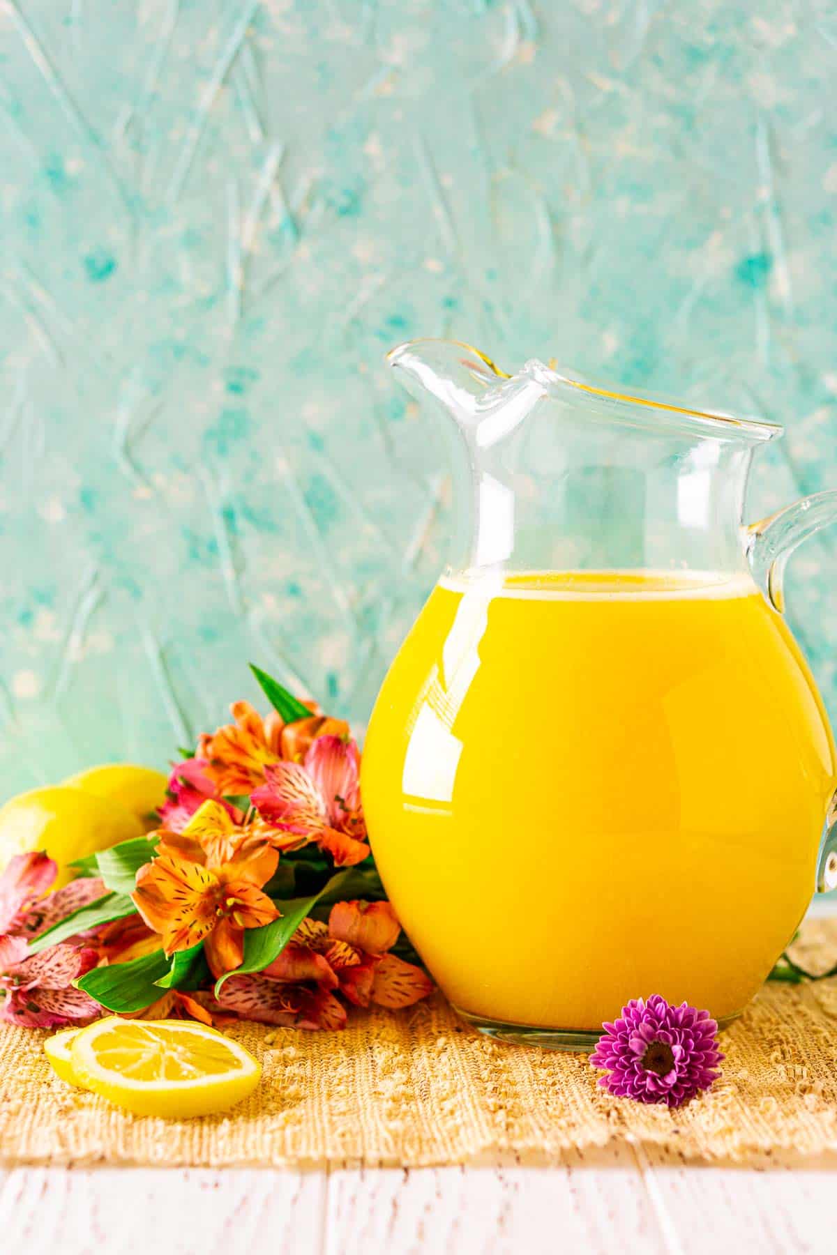 A glass pitcher filled with the mango lemonade against a blue background on a straw placemat with tropical flowers around it.