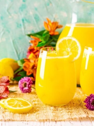Looking straight on to a glass of mango lemonade on a straw placemat with colorful flowers around it.
