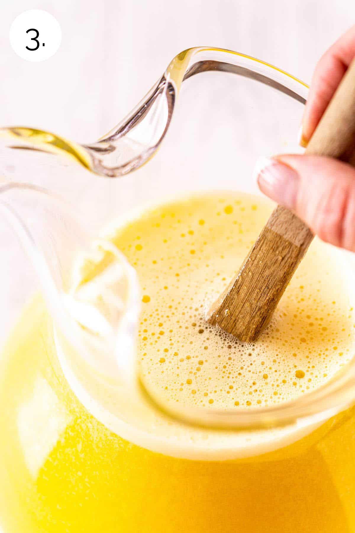 A hand stirring a brown wooden spoon in a glass pitcher to combine the ingredients and make the lemonade.
