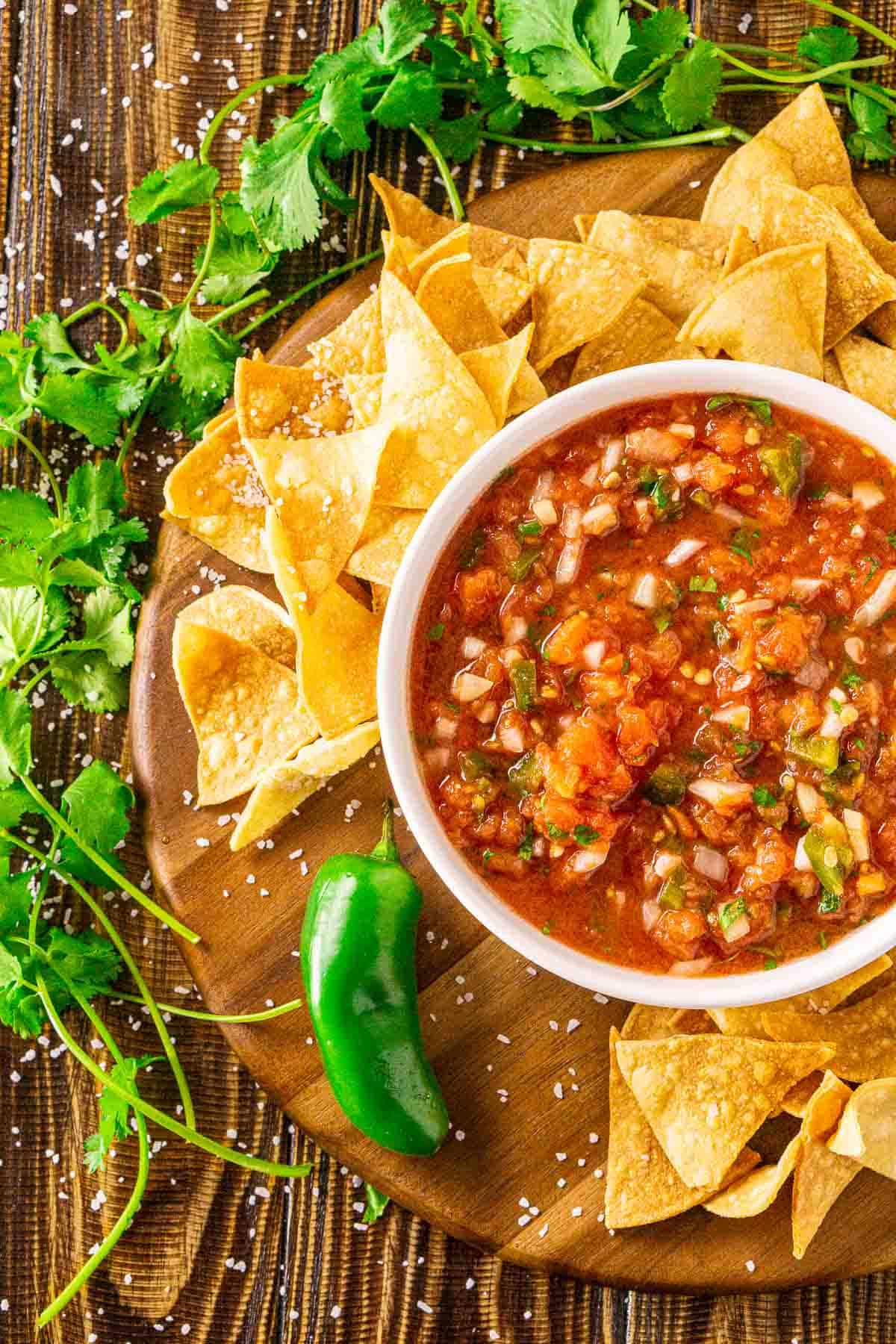 An aerial view of a white bowl filled with the smoked salsa with sprigs of cilantro and a jalapeño pepper to the left and chips scattered around it.