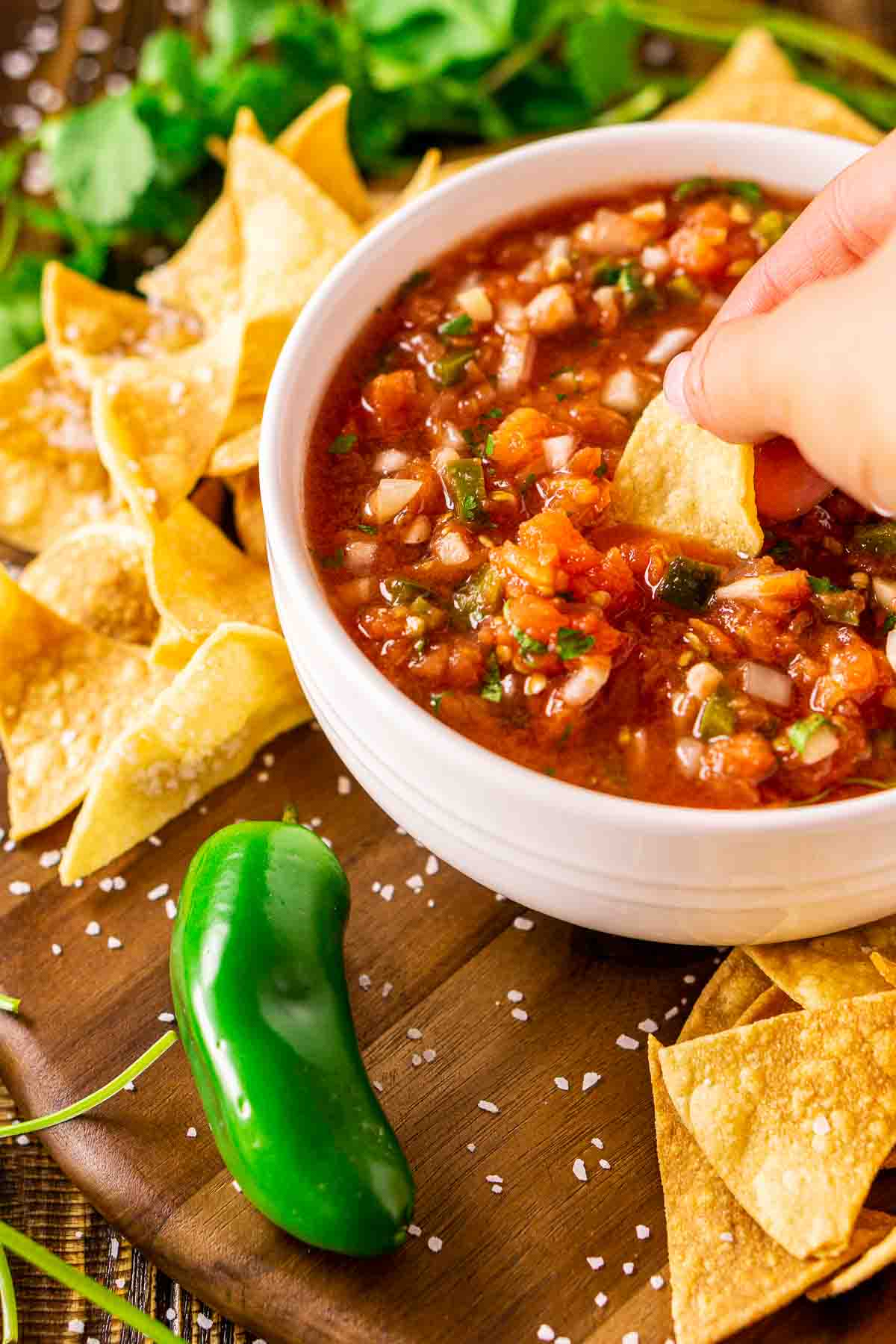 A hand dipping a tortilla chip into a white bowl of the smoked salsa with a jalapeño to the bottom left.