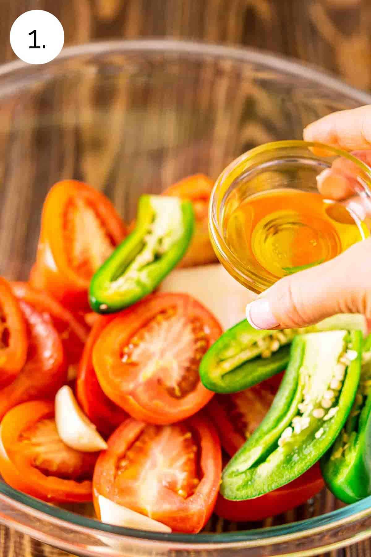 A hand pouring a small bowl of olive oil over the vegetables in a large glass mixing bowl.